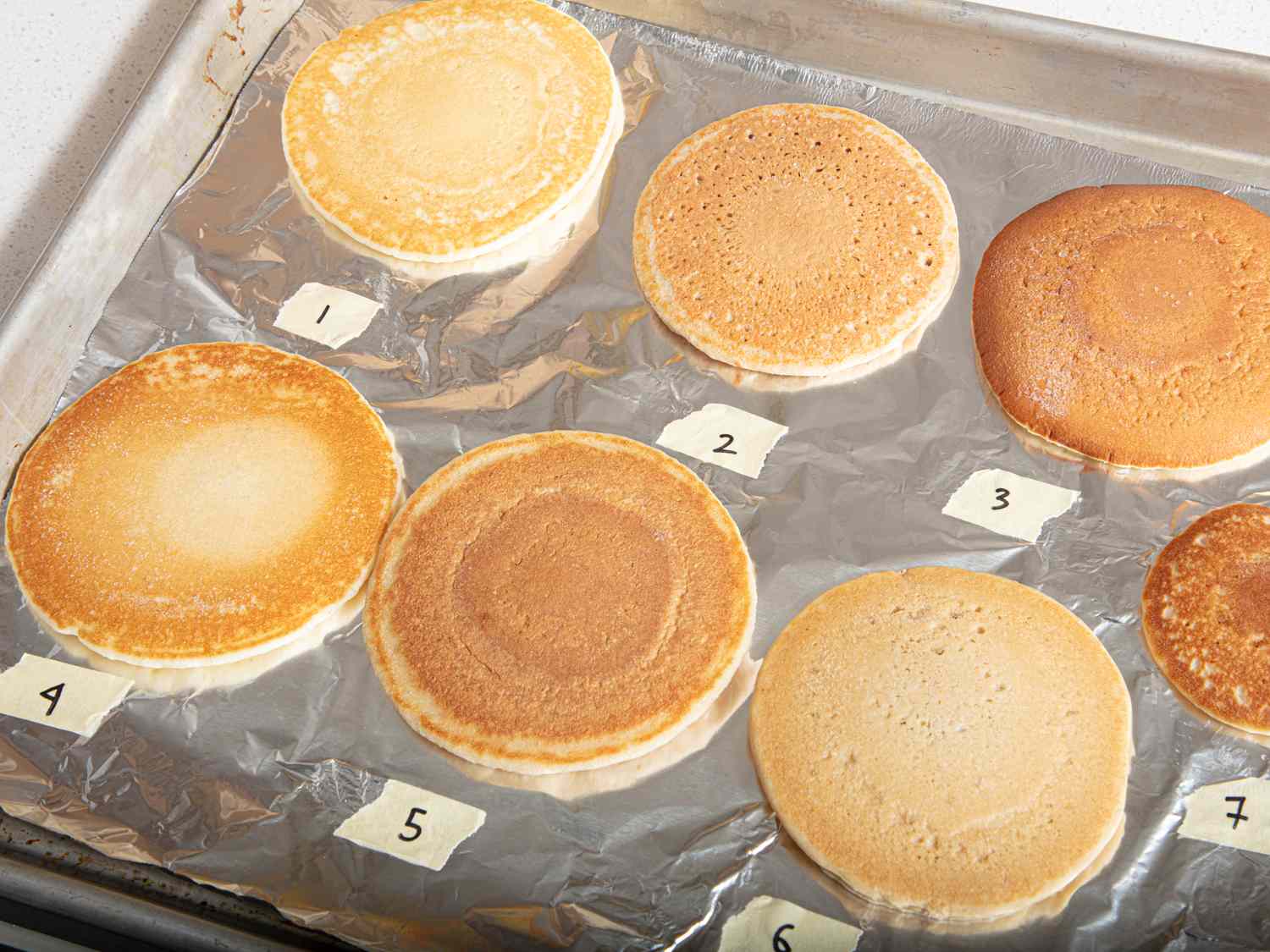 Seven cooked pancakes labeled with numbers on a foillined baking tray