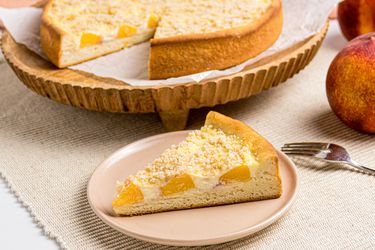 A slice of peach streusel cake on a plate with a larger cake in the background on a wooden stand
