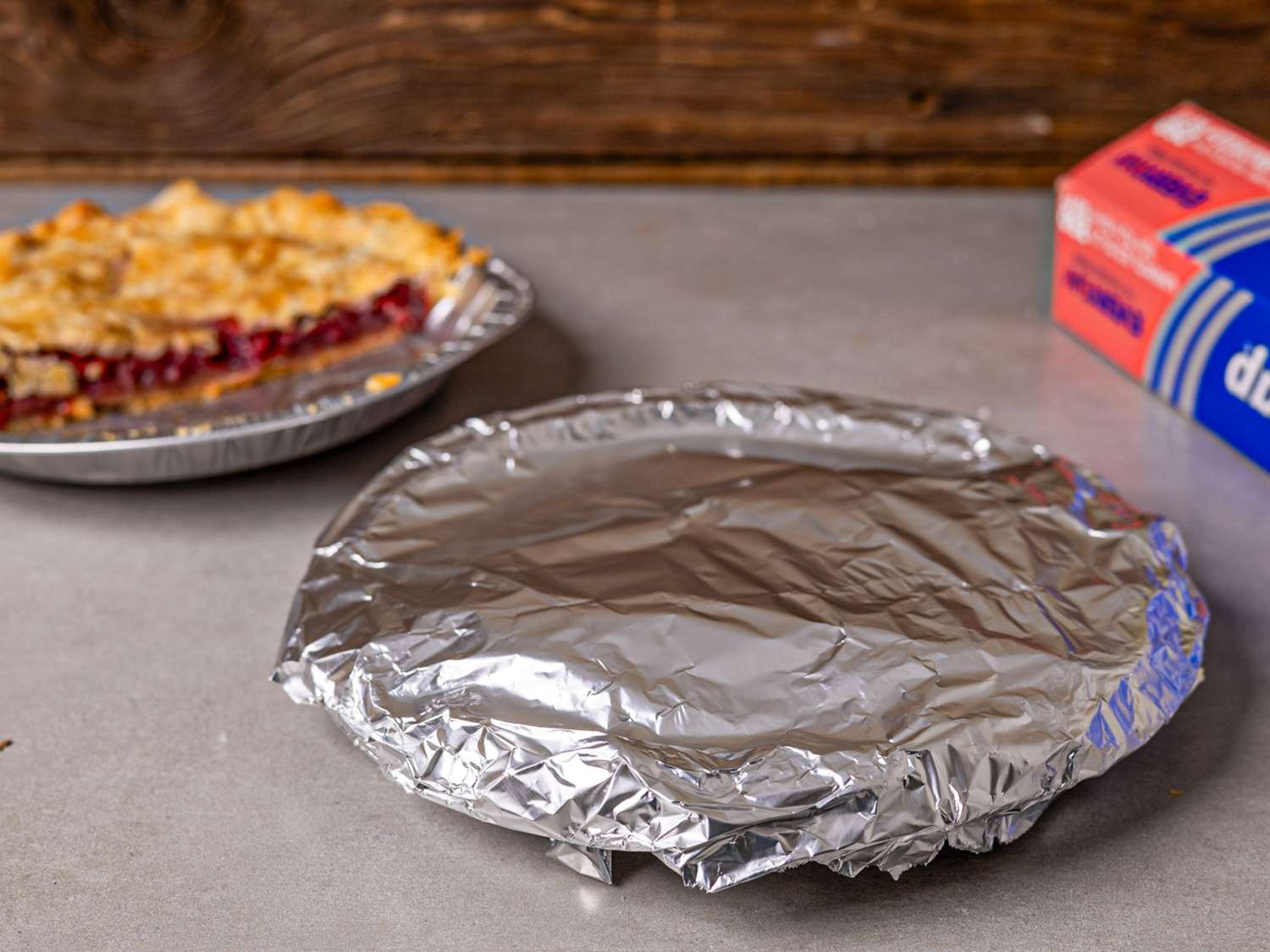 A pie covered in aluminum foil on a countertop another uncovered pie and a box of foil in the background