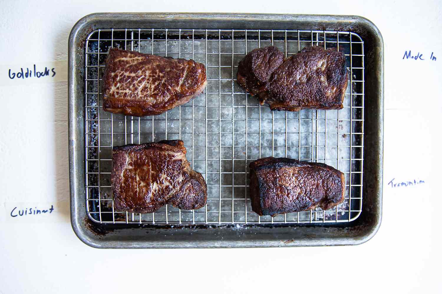 Four cooked pieces of meat on a wire rack with labels Goldilocks, Made In, Cuisinart, and Tramontina written on the sides
