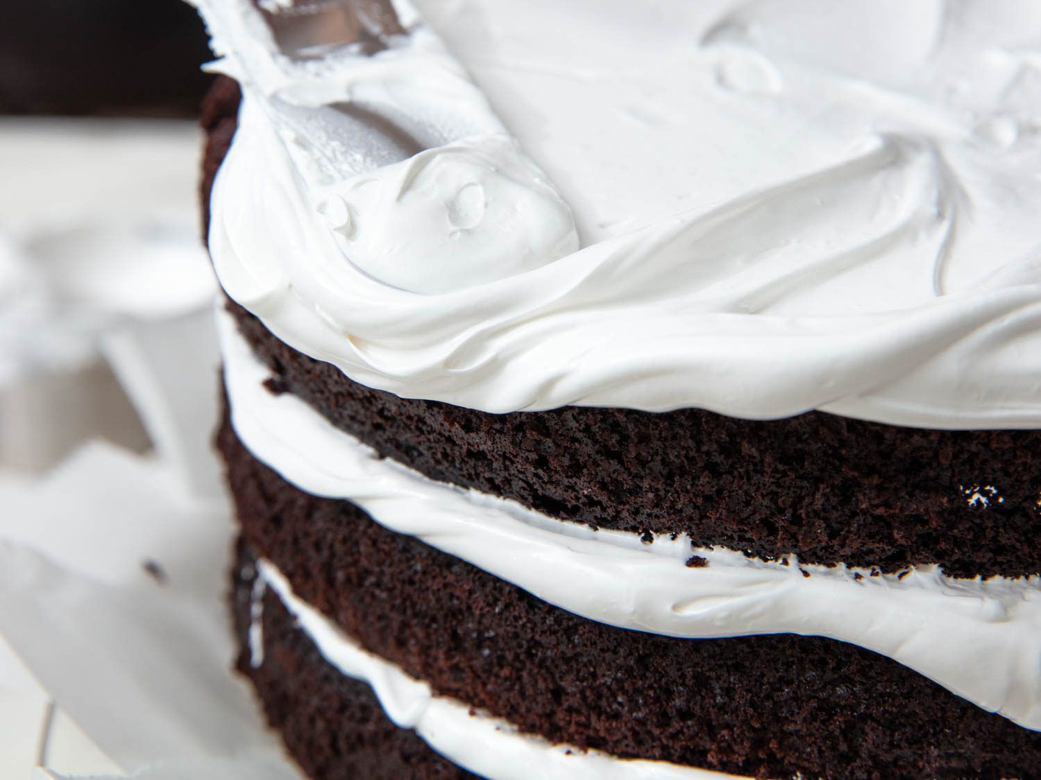 an offset spatula spreads fluffy 7-minute frosting over the top of a three layer chocolate cake, its dark sides still exposed