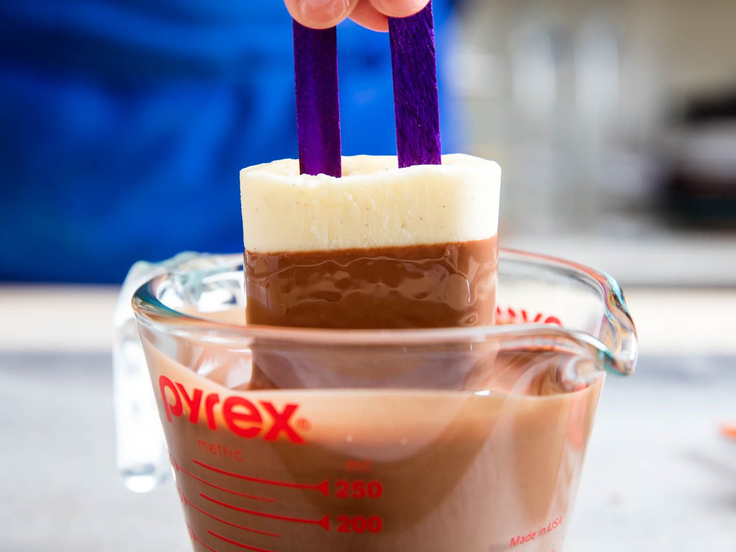 A pudding pop is dipped into a liquid measuring cup full of chocolate magic shell.
