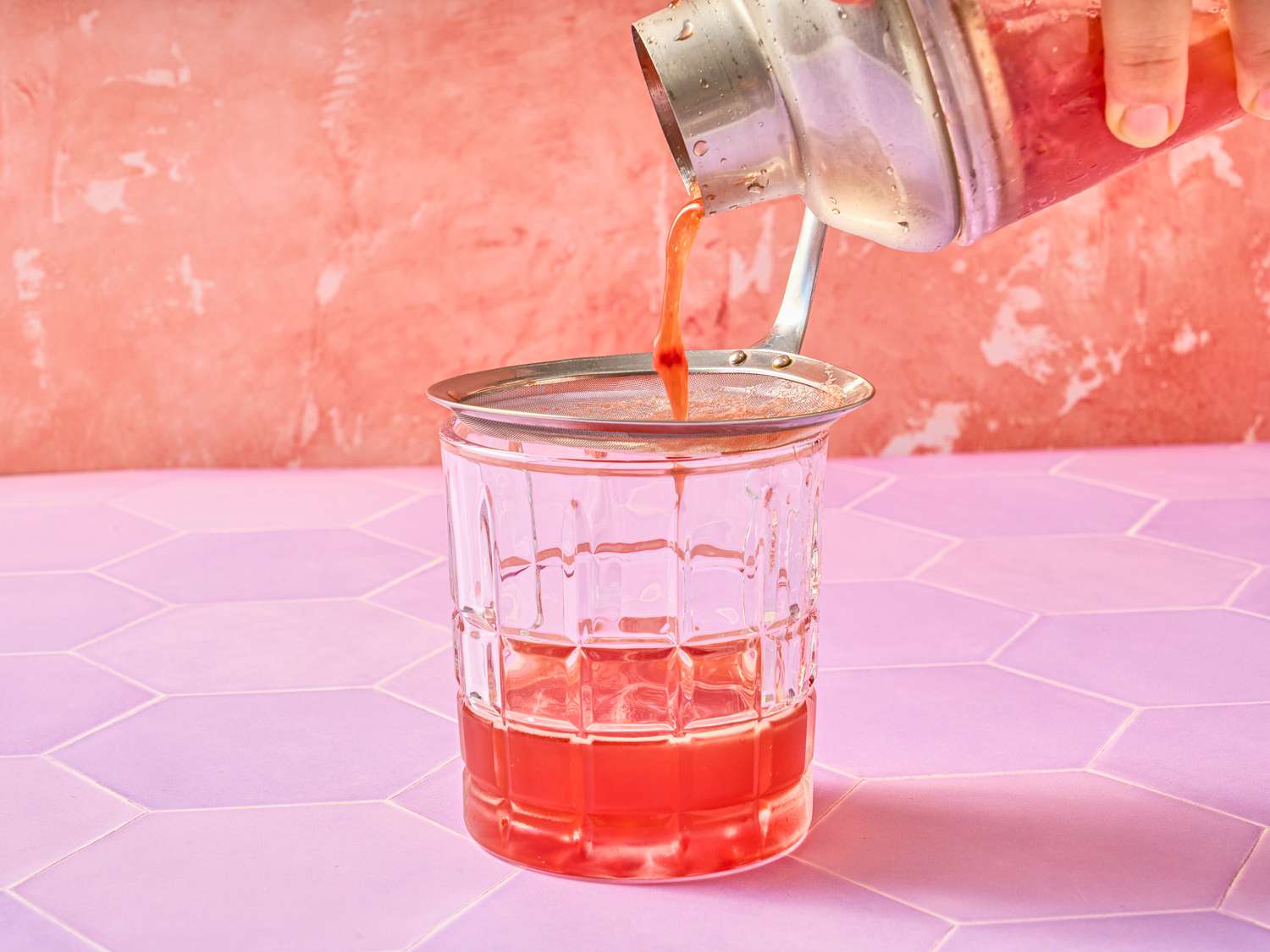 A cocktail being poured through a strainer into a glass on a pink surface