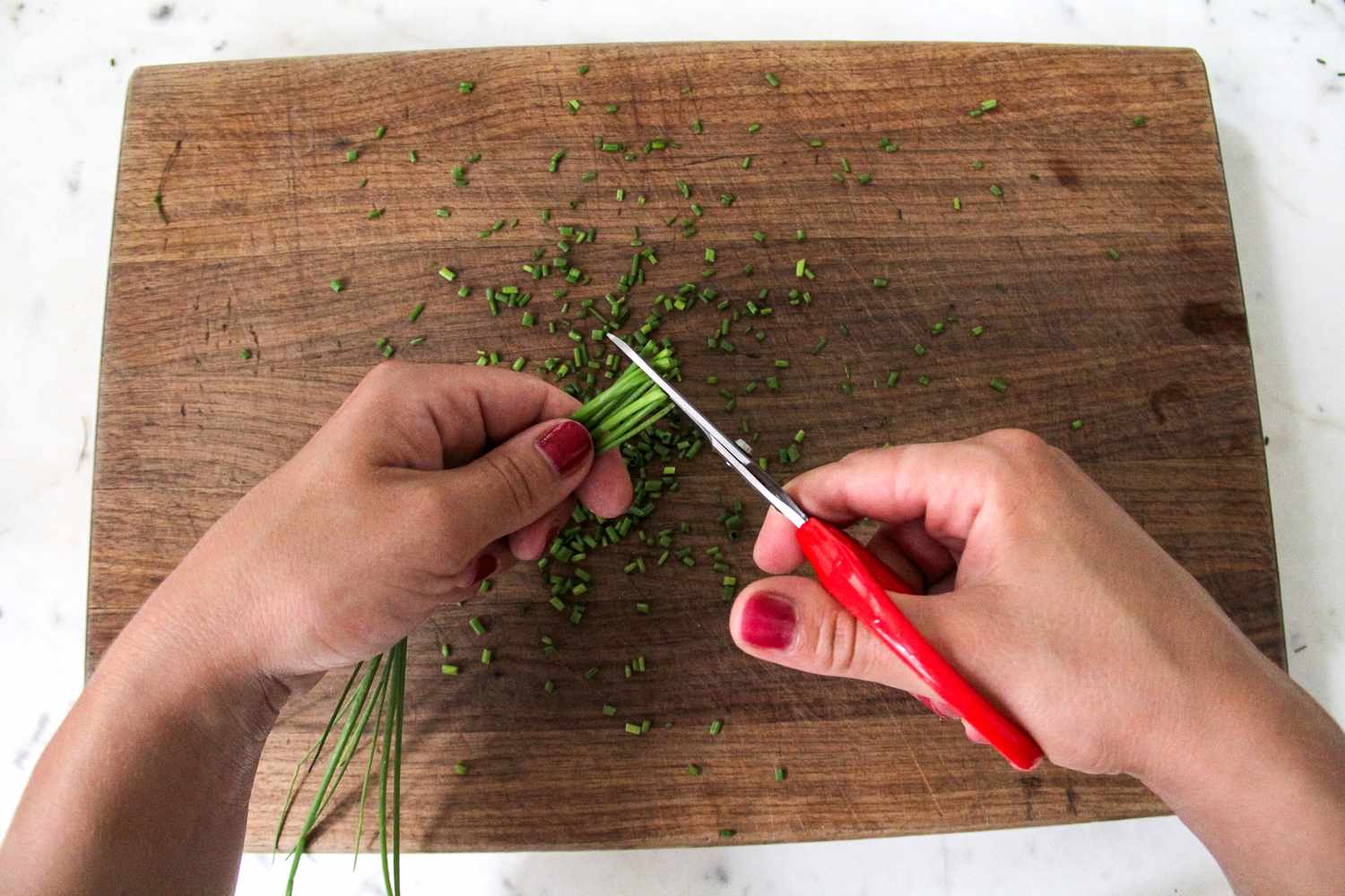 Hands using kitchen scissors to cut chives on a wooden cutting board