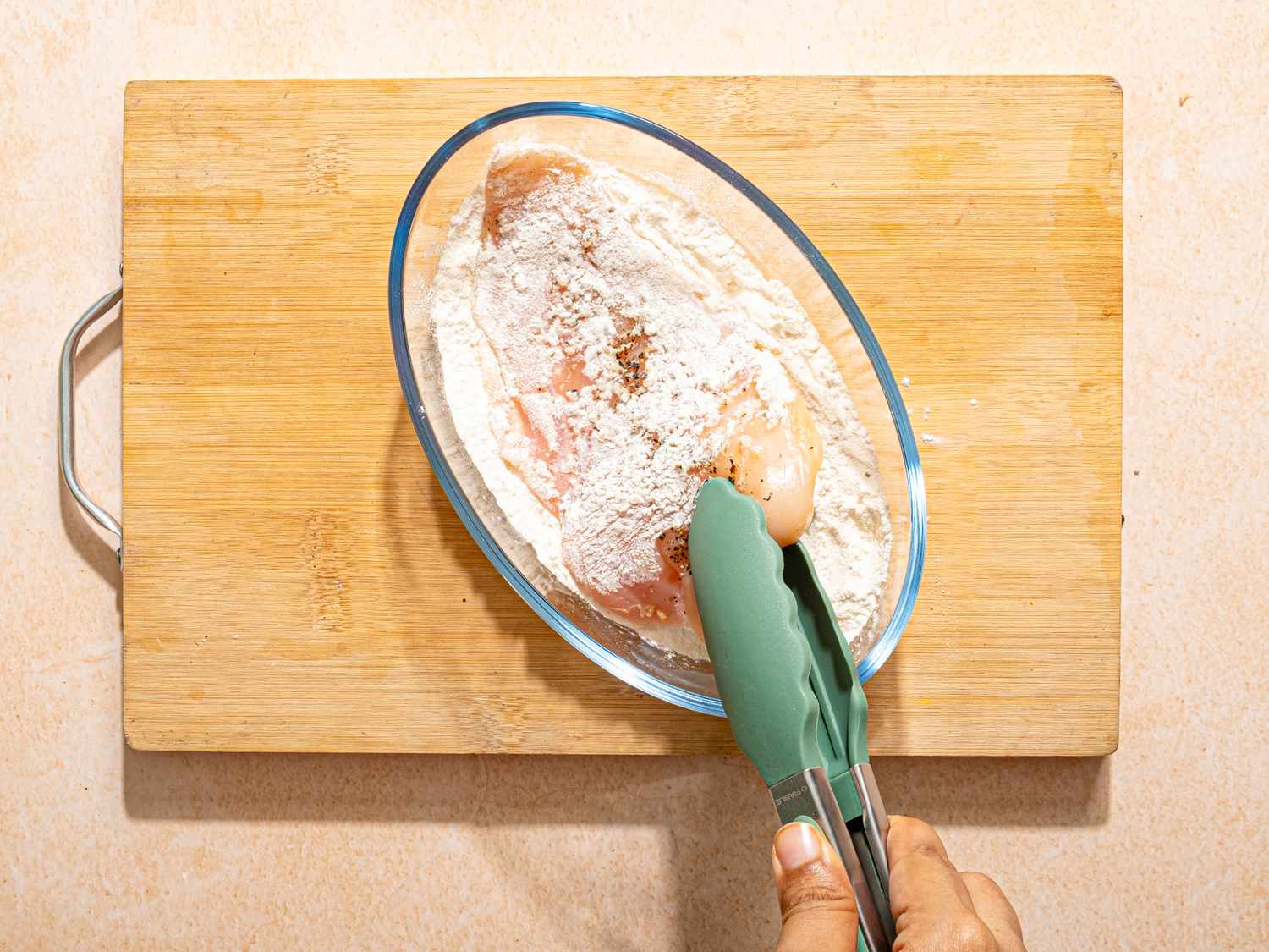 A person using tongs to coat chicken breasts in flour on a cutting board