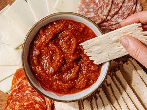 A hand holding a cracker above a bowl of red dip surrounded by slices of cheese and meats