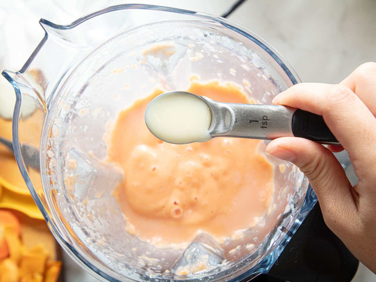 A hand pouring a small amount of liquid from a teaspoon into a blender containing a fruit mixture