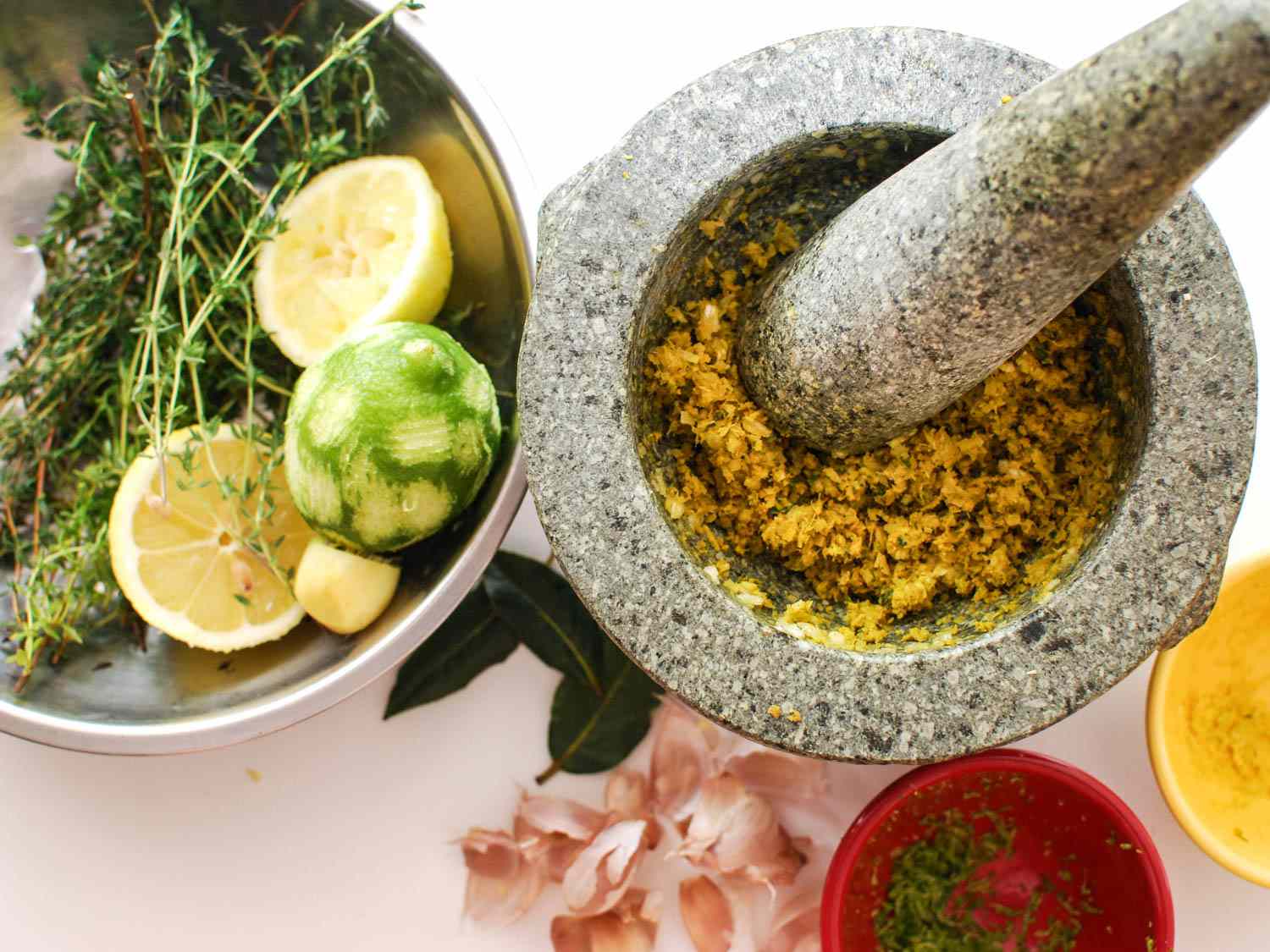 Aromatics pounded into a paste with mortar and pestle. Next to the mortar is a mixing bowl containing lime, lemon, and thyme.