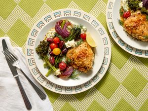 A plate of roasted chicken with vegetables and feta on a patterned plate on a green background additional plate and utensils visible