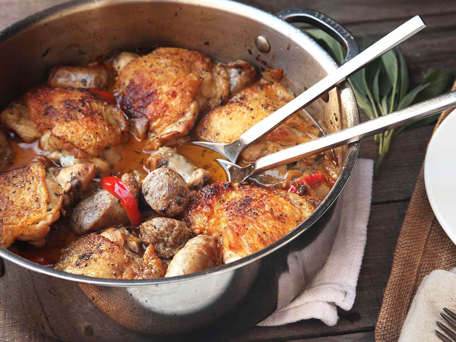 Braised chicken with sausage and red pepper in a stainless steel skillet with serving utensils.