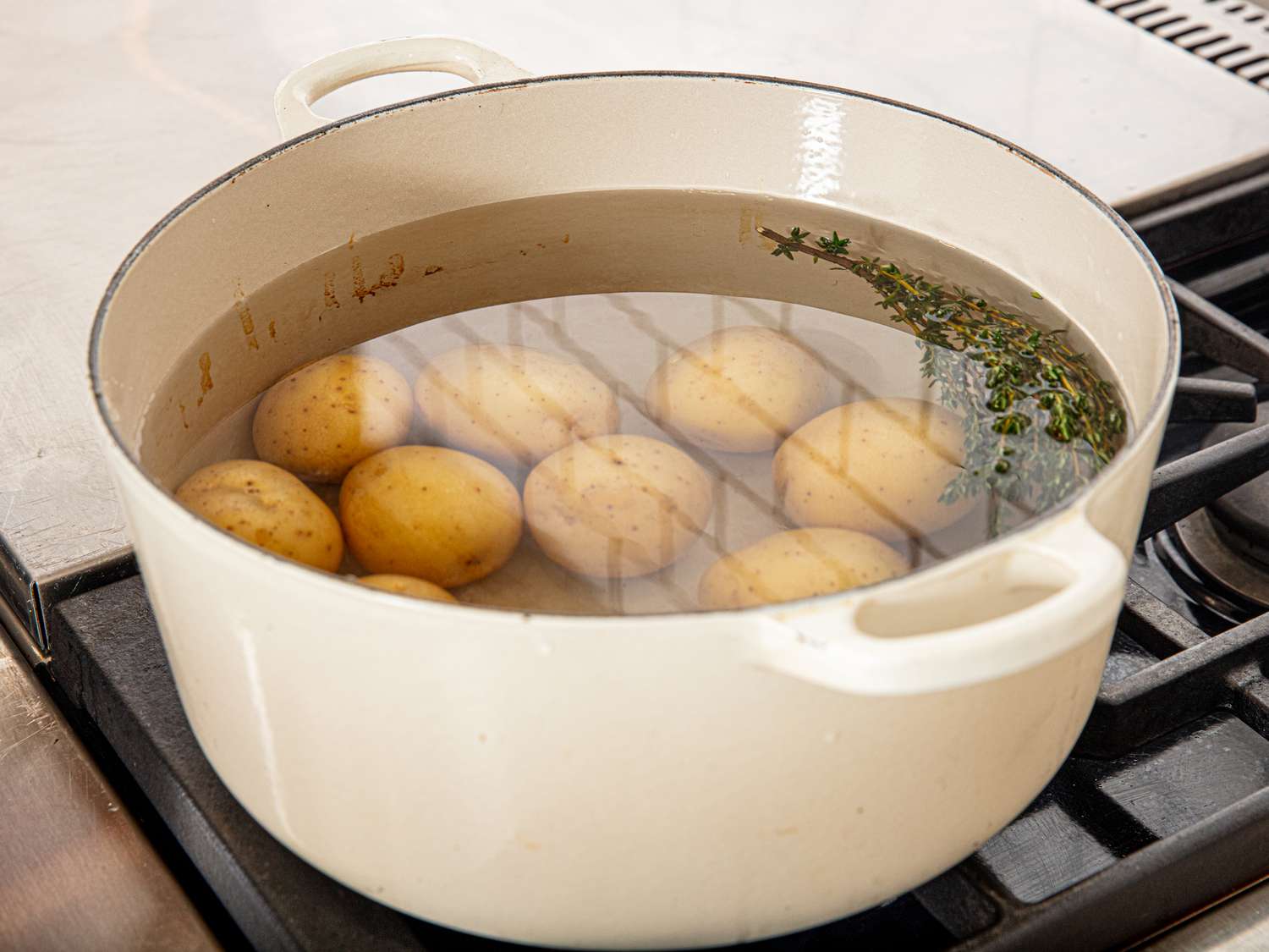 Potatoes boiling in a white pot with sprigs of herbs on a stove