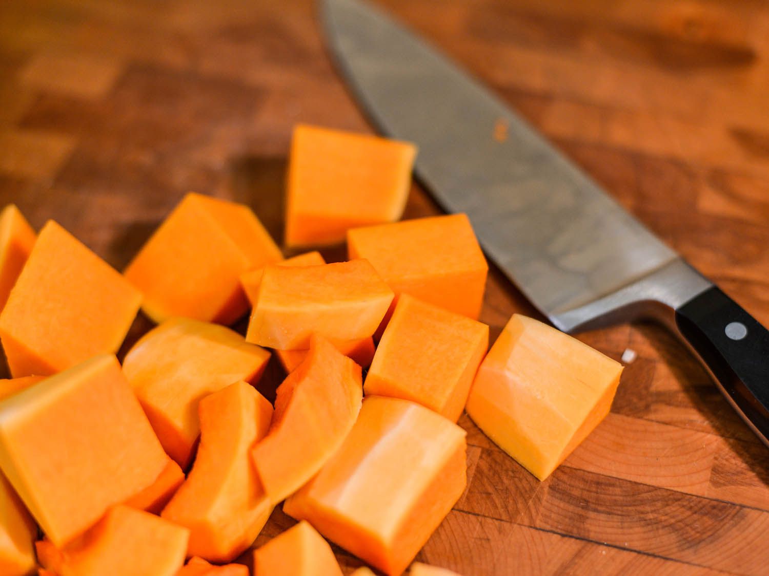 Butternut squash cubes on a wooden cutting board, lying next to a chef's knife.