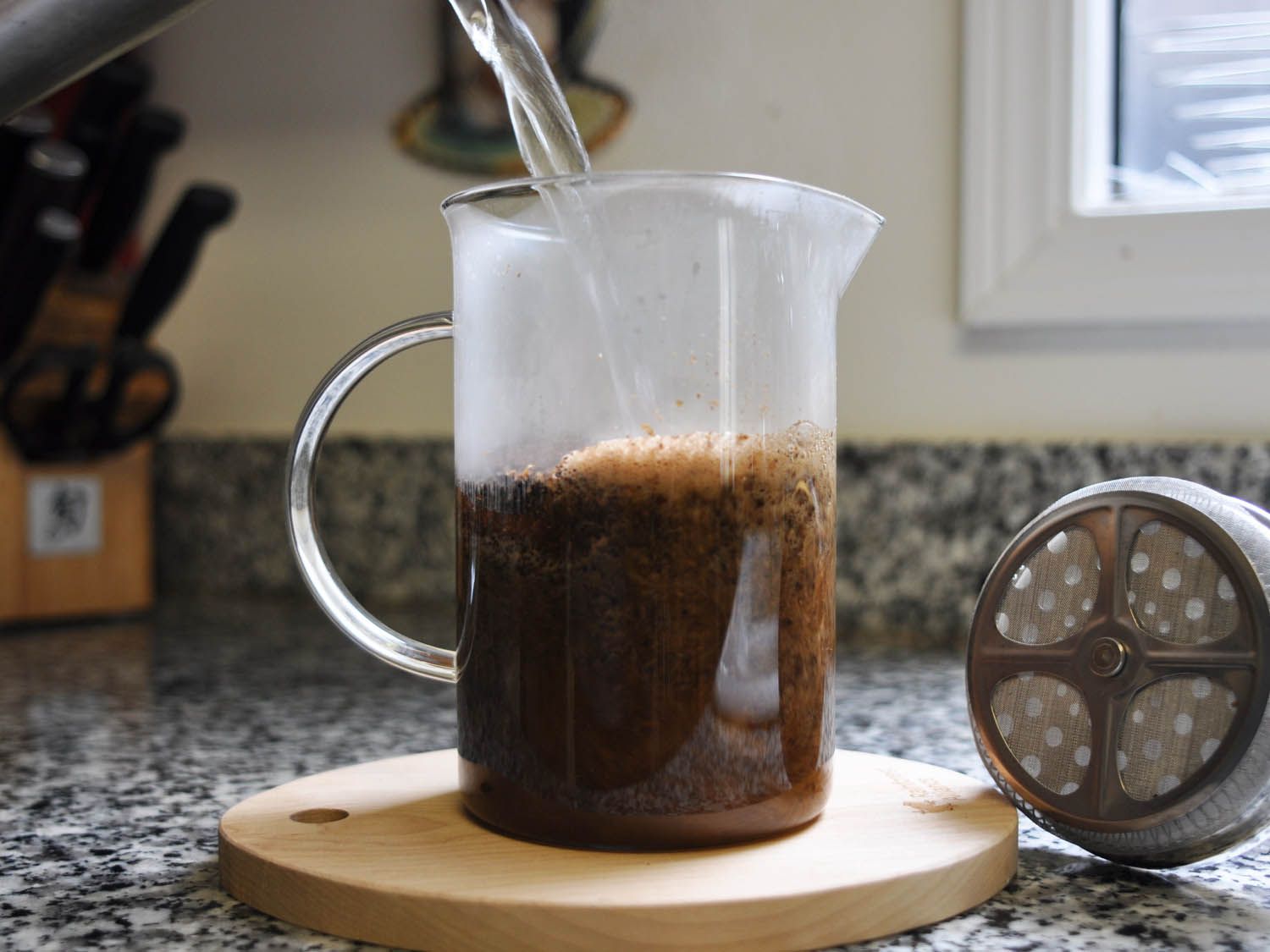 Hot water being poured into a glass French press with ground coffee.