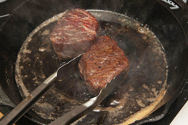 Two small beef steaks cooking in a cast iron skillet. A pair of tongs is lifting one of the steaks. 