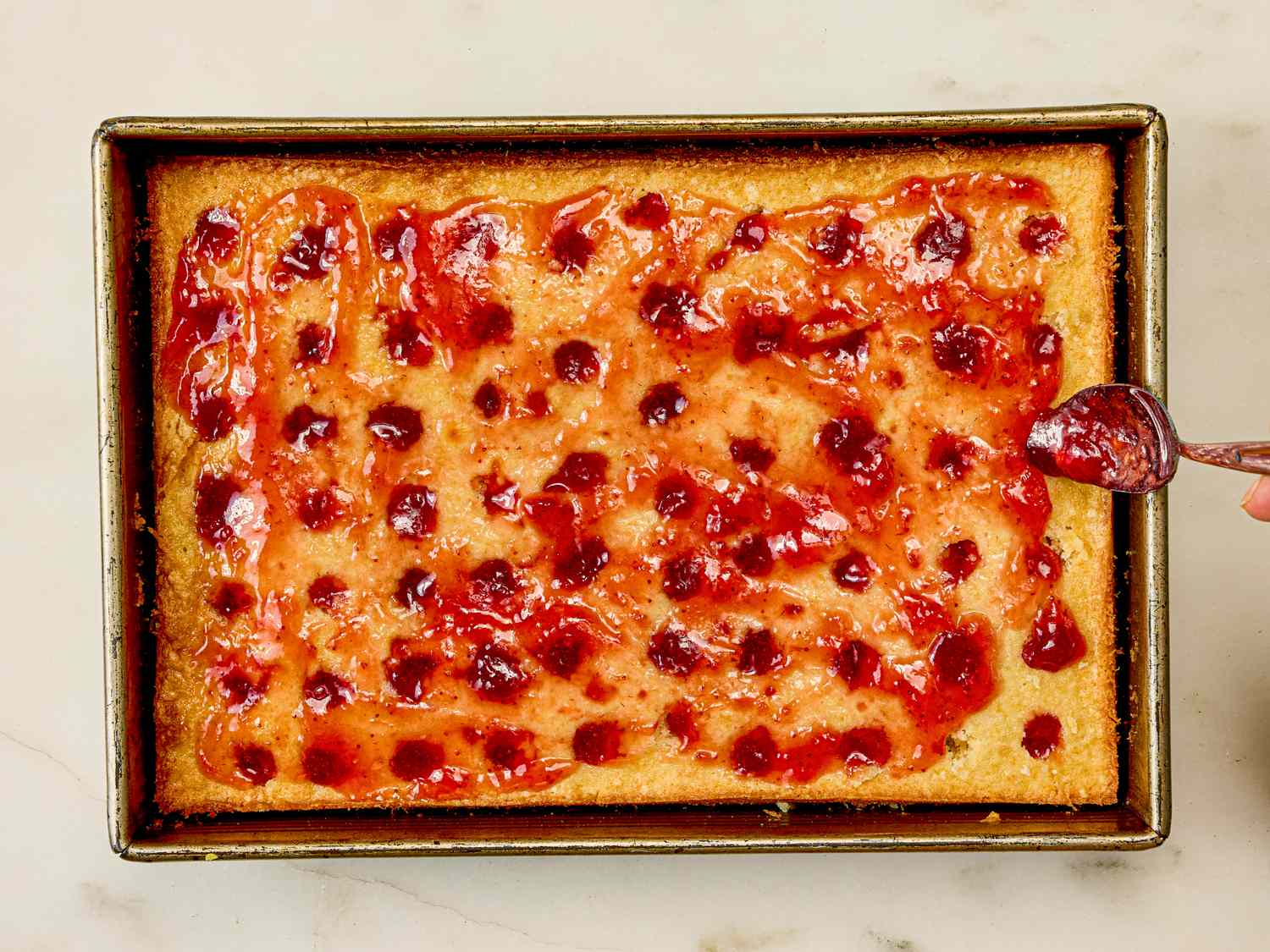 A baking pan containing a strawberry poke cake with a spoon spreading strawberry topping
