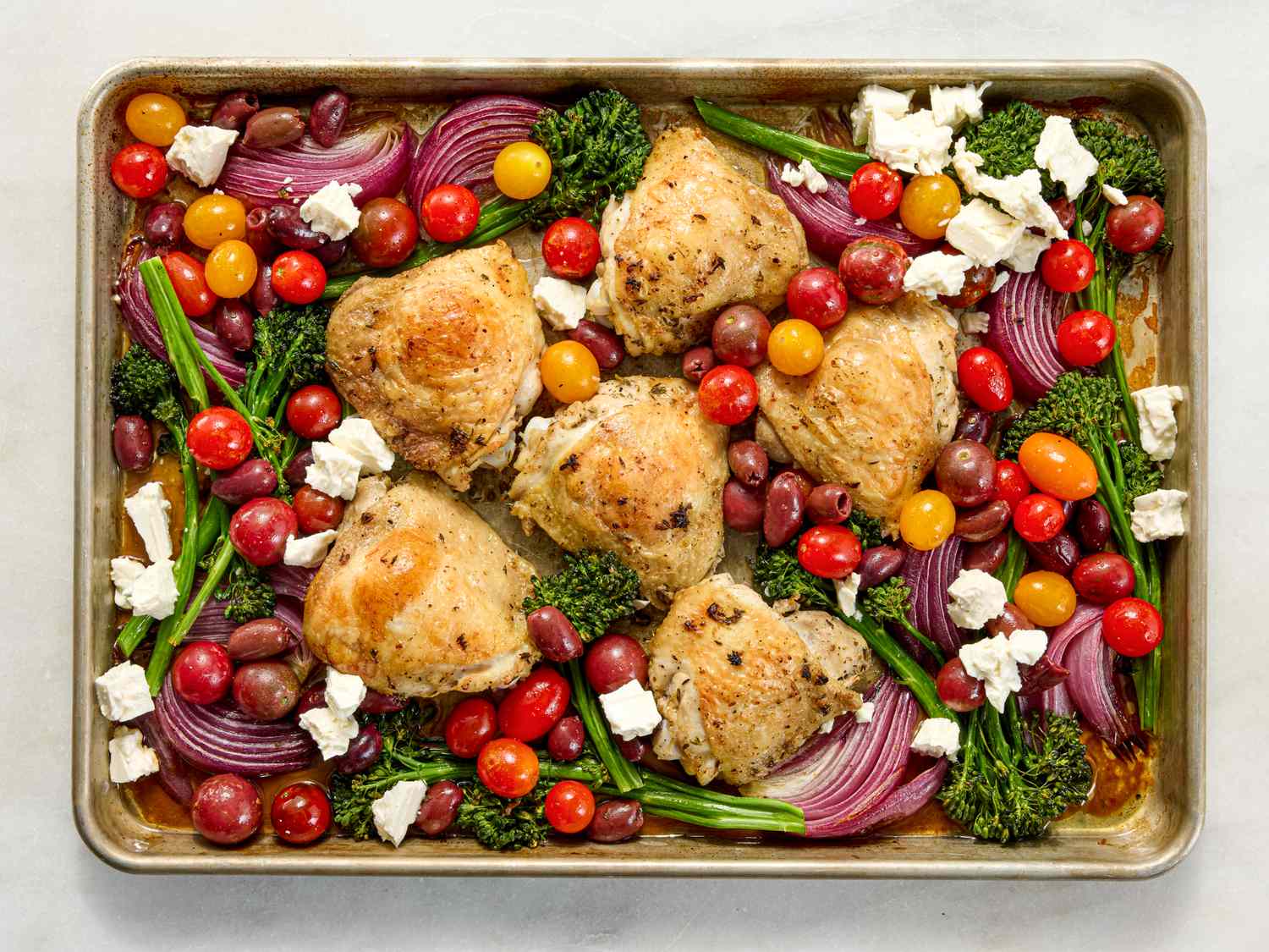 Sheet pan with roasted chicken thighs tomatoes red onions olives broccolini and feta cheese crumbles