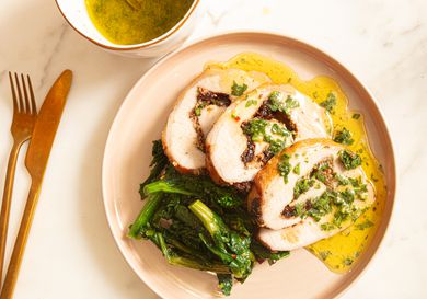 Stuffed pork loin slices with sauce sauce and greens on a warm colored plate. Gold fork and knife to the side and small bowl of sauce in top corner 