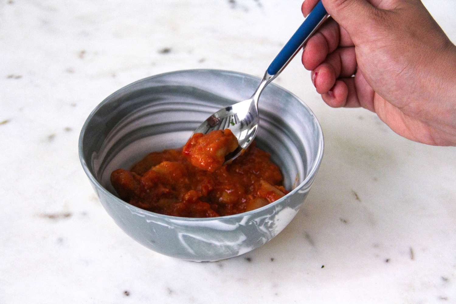 A hand holding a spoon with a portion of food from a bowl of stewlike dish