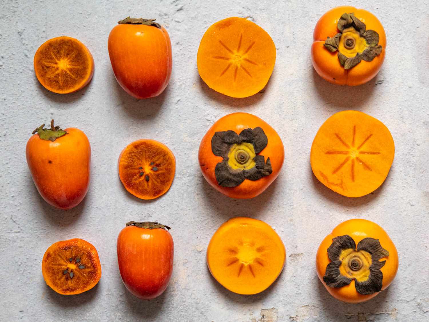 A variety of persimmons, some cut and some whole, arranged in a grid on a stone surface
