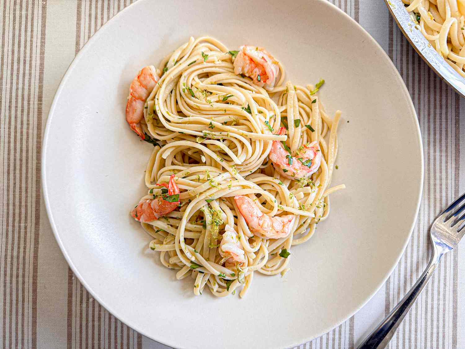A plate with spaghetti and shrimp, garnished with herbs, placed on a table with a fork nearby