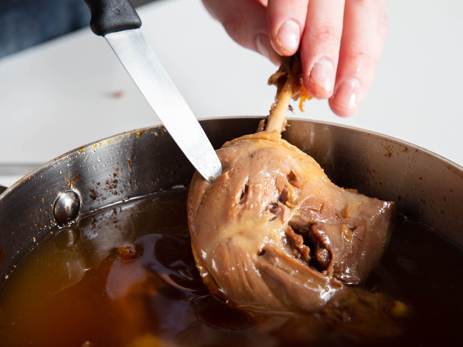 The finished duck confit is held above the duck fat and prodded with a paring knife to ensure it is fall-off-the-bone tender.