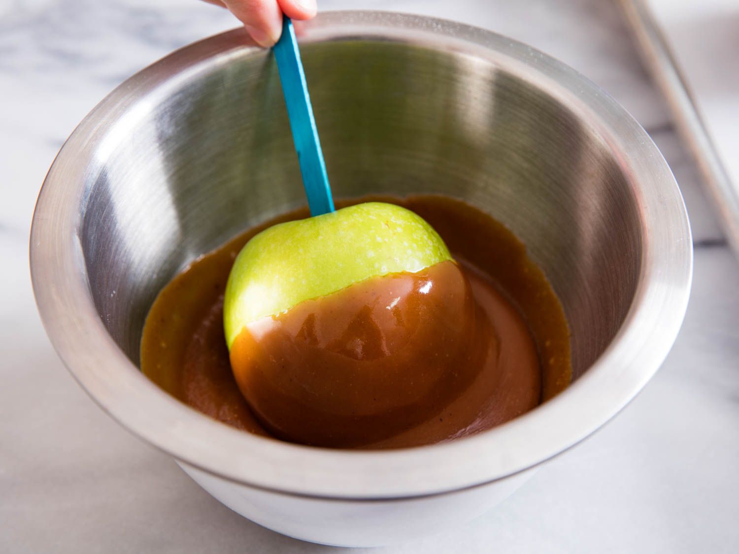 Using a blue popsicle stick as a handle to dip a green apple into a metal bowl of caramel.