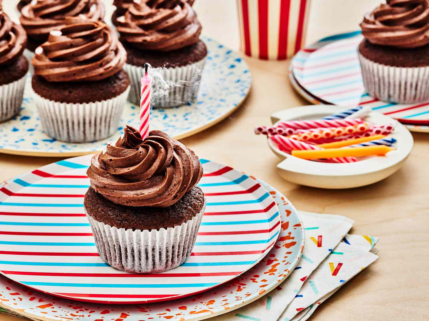 Chocolate cupcake with smoking candle in the frosting on a stack of colorful plates. In the background, there is a plate of more cupcakes and a bowl of birthday candles. 