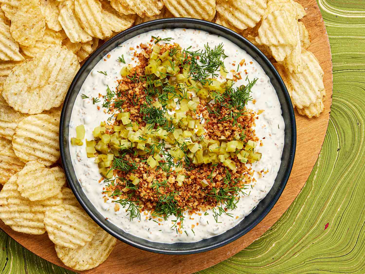 Bowl of pickle dip garnished with panko, dill and pickles, surrounded by a wooden plate full of potato chips, on a green tabeltop