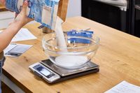 A person pours white sugar into a glass bowl on the OXO Good Grips Stainless Steel Food Scale with Pull-Out Display