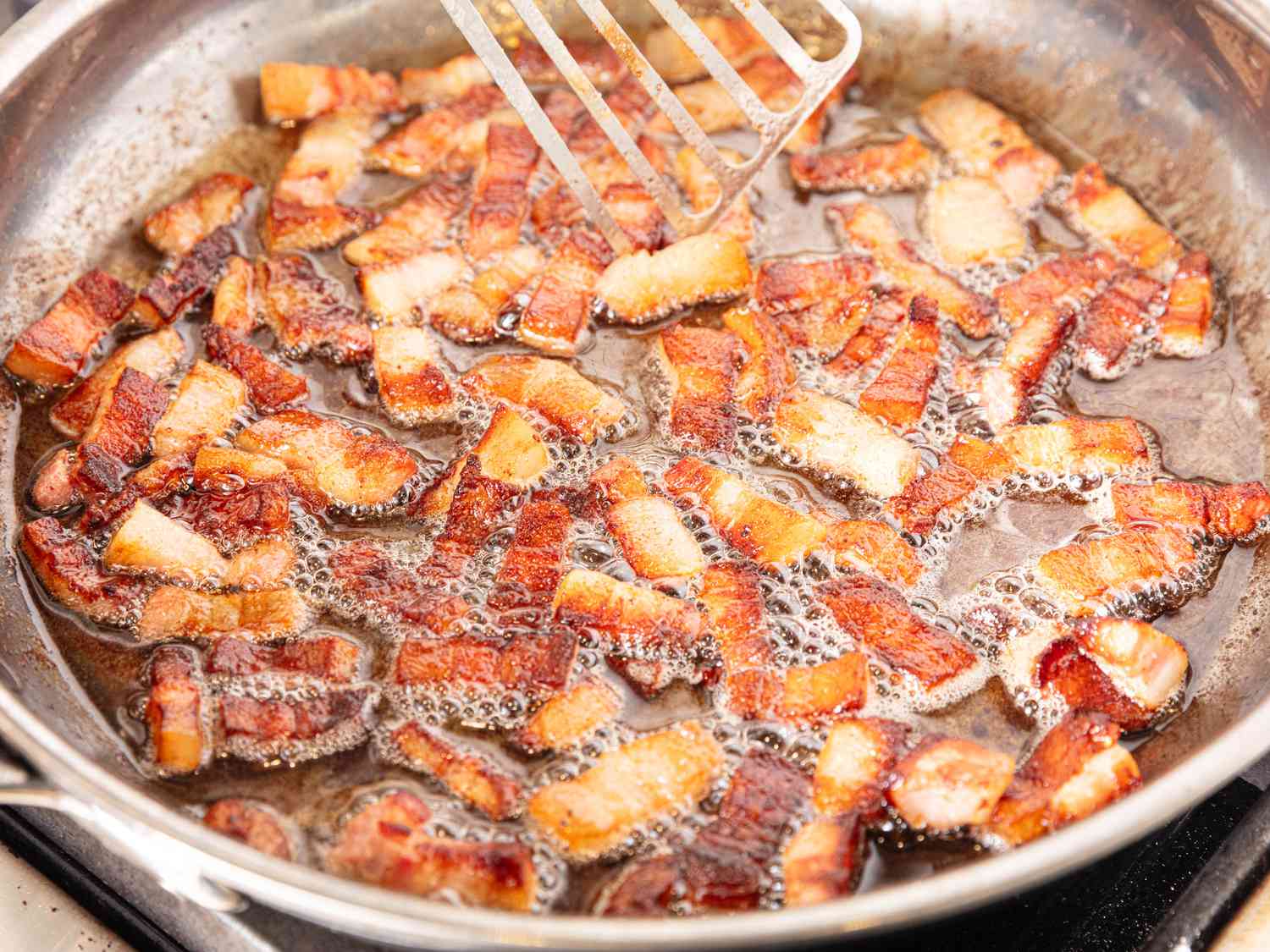 Pancetta crisping in oil on a skillet with a slotted spatula 