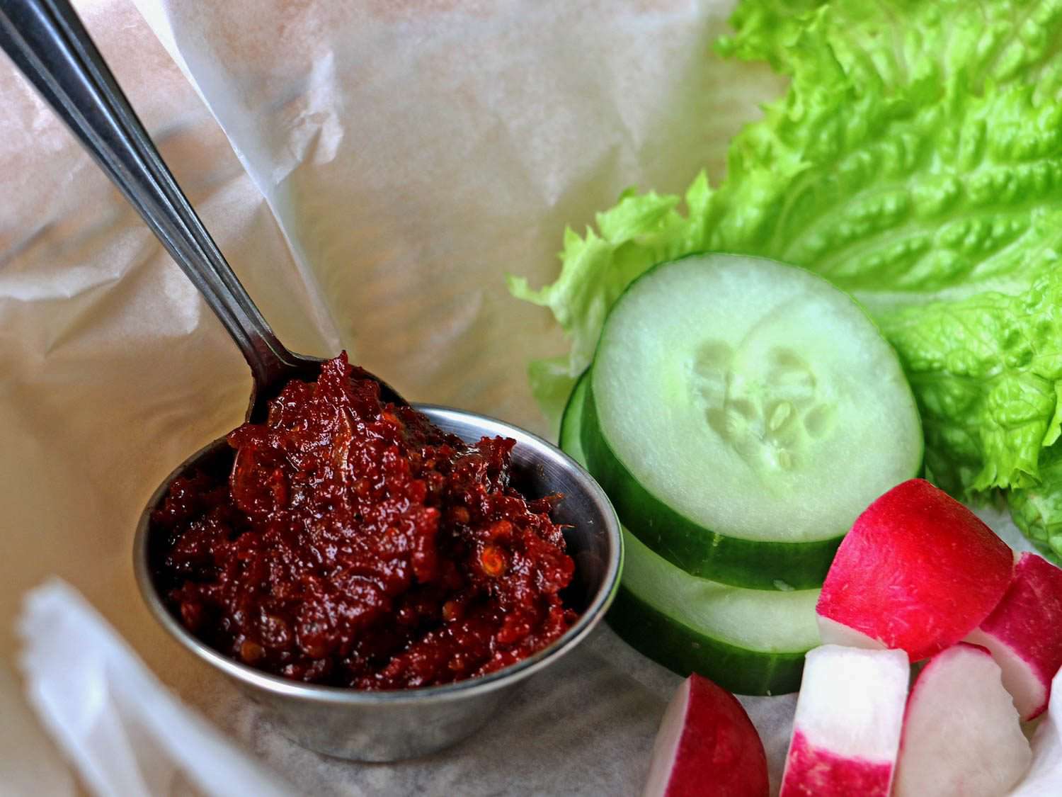 A small silver bowl of dark red hot pepper dip (jaew bong) with a spoon dipped into it, next to raw cucumber, radish, and lettuce.