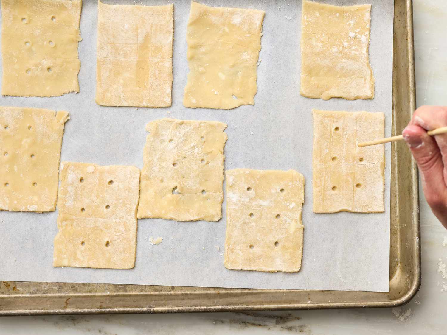 Rectangular pastry sheets pierced with holes on a parchment-lined baking tray, a hand holding a wooden skewer nearby