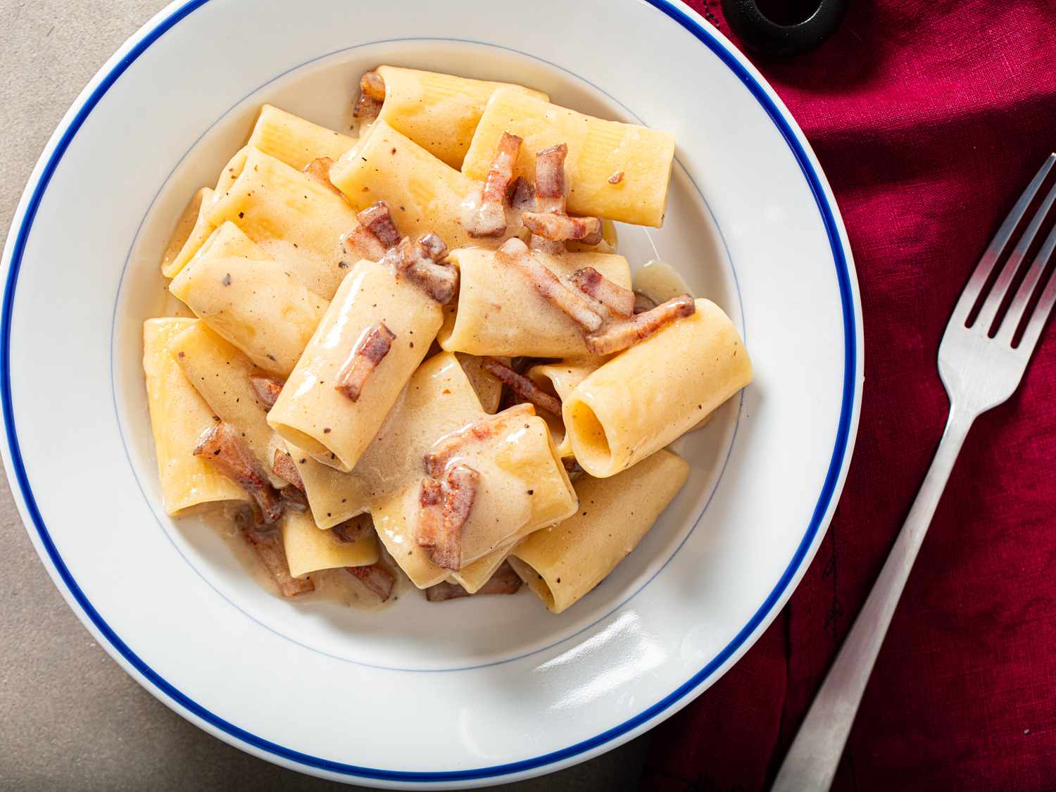 A bowl of pasta served with a creamy sauce and small pieces of meat fork and napkin beside the dish