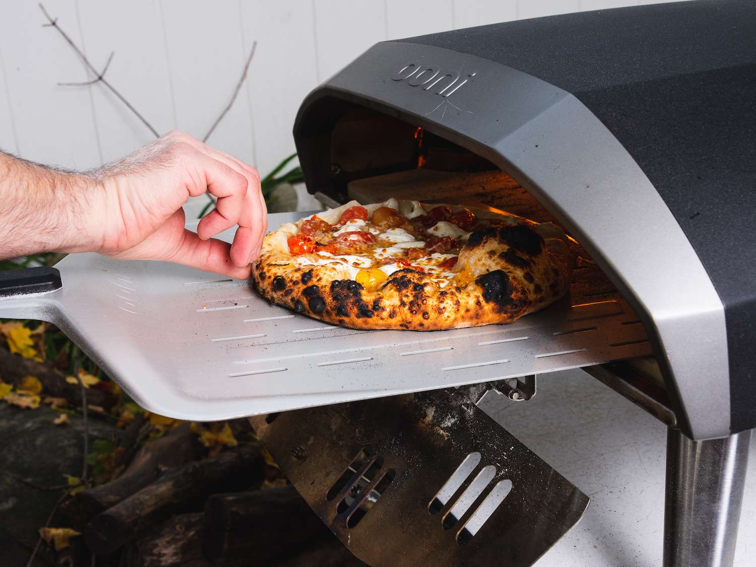 Rotating a pizza with burst cherry tomatoes as it bakes in an outdoor pizza oven
