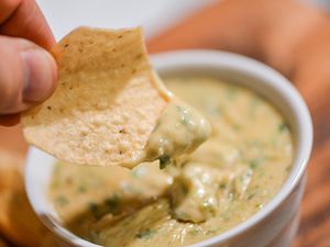 A tortilla chip dipped in green chile queso is held up to the camera.