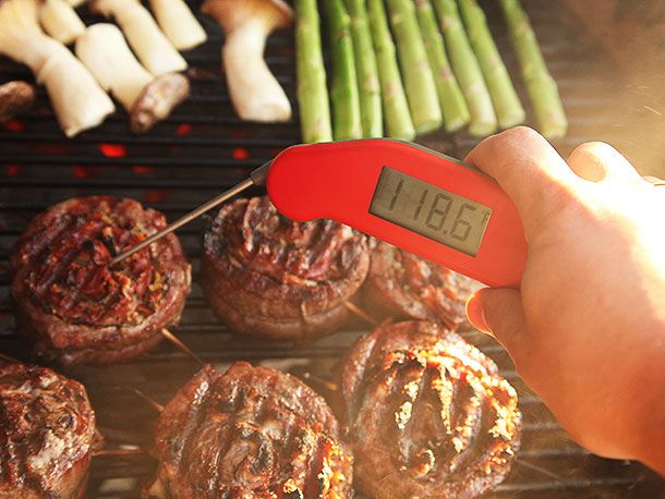 Hand holding a digital thermometer that's testing temperature of a stuffed flank steak on barbecue