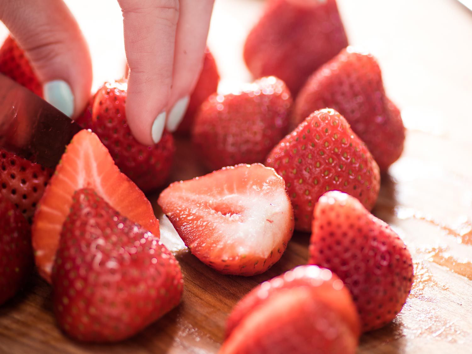 A hand holding a strawberry steady while cutting it in half, next to a pile of other strawberries on a wooden surface