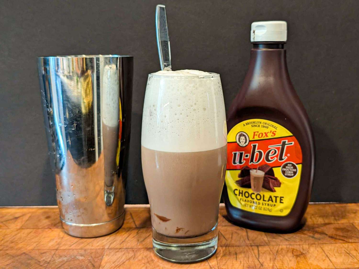 A chocolate egg cream in a glass with a spoon next to a metal shaker and a bottle of Foxs Ubet chocolate syrup