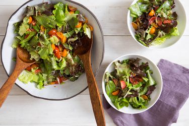 Overhead view of a green salad with tomatoes, scallions, and toasted kasha being served from a large off-white salad bowl with wooden serving utensils.