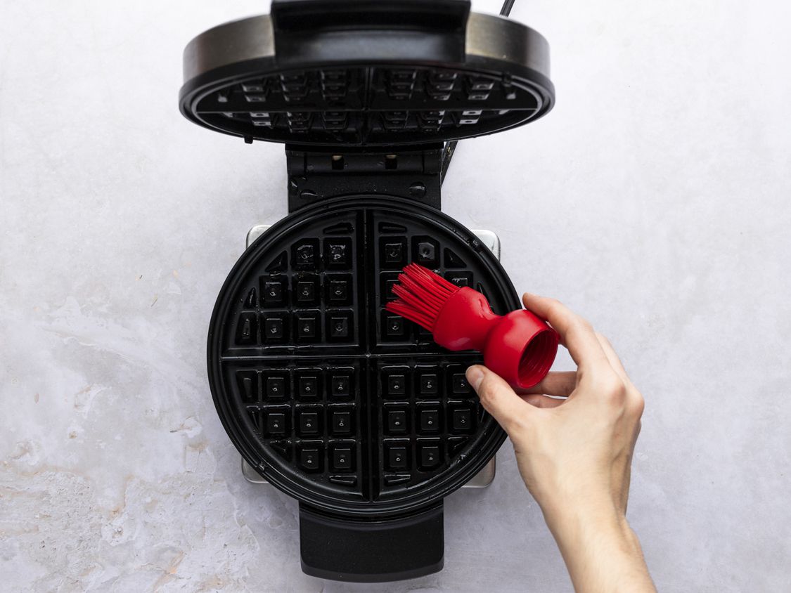 The open waffle iron being greased with butter with a red silicone brush held by a hand.