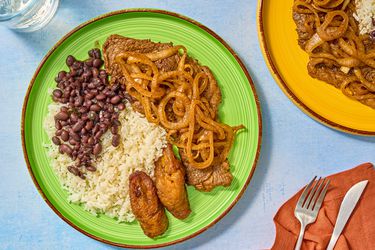 Green Plate of Pallomilla with rice beans and plantains. Red napkin with untensils, and large plate of pallomilla to the side 