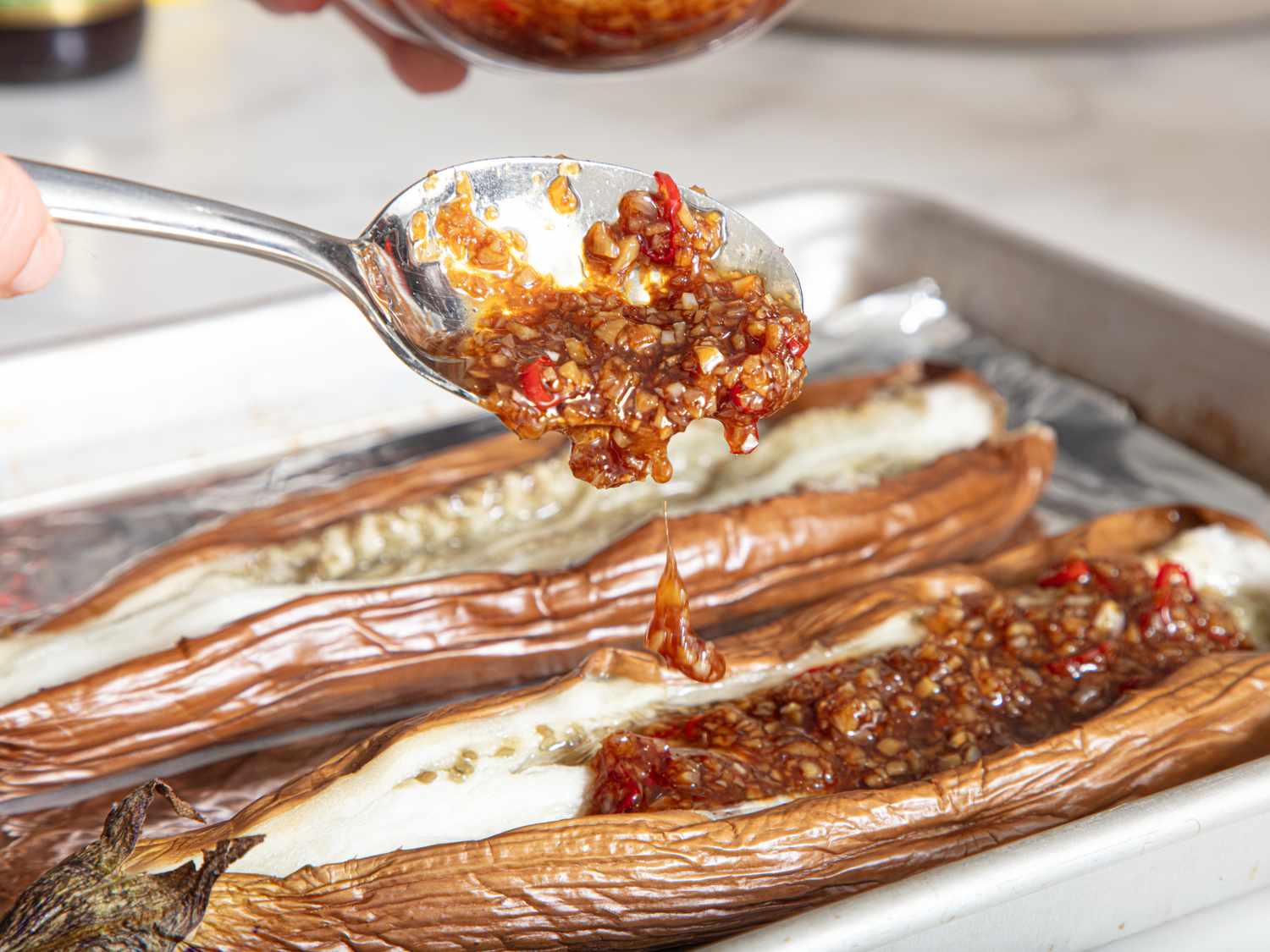 Roasted eggplants topped with a flavorful sauce being applied with a spoon on a baking tray
