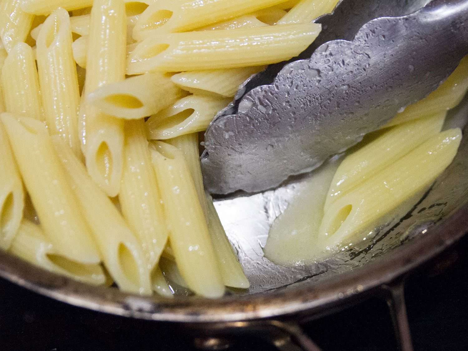 A pair of metal tongs moving cooked penne around in a pan, with starchy water clearly evident.