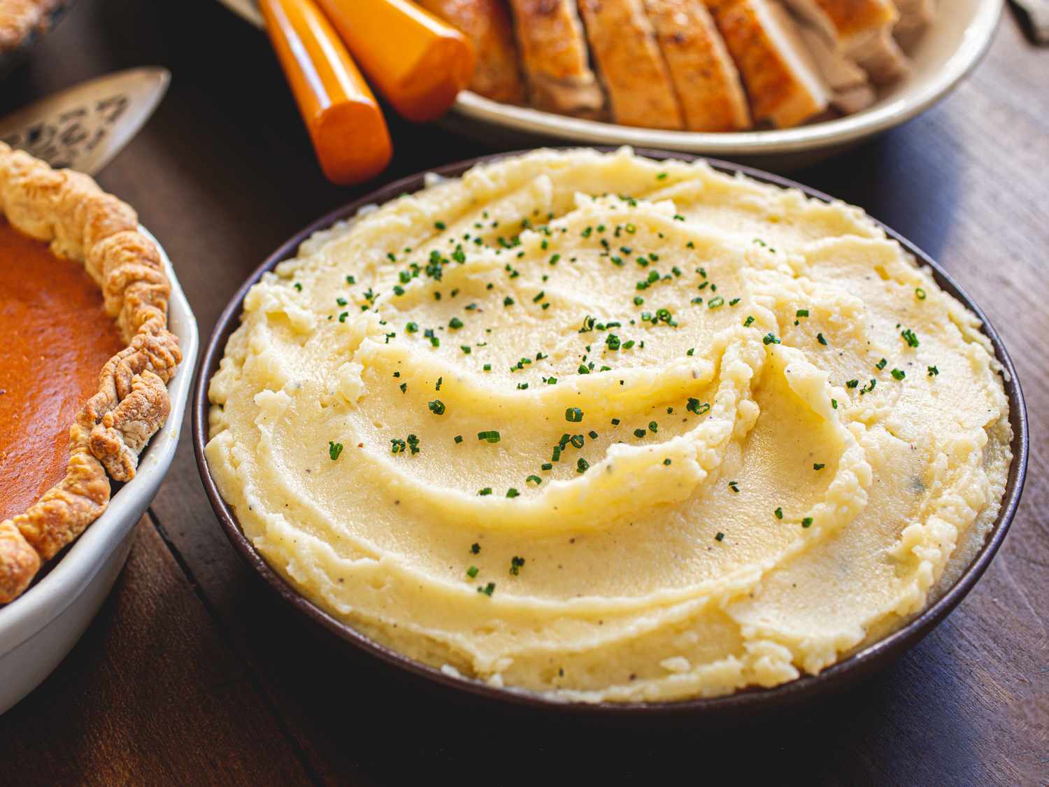 A bowl of mashed potatoes garnished with chives on a table with Thanksgiving dishes