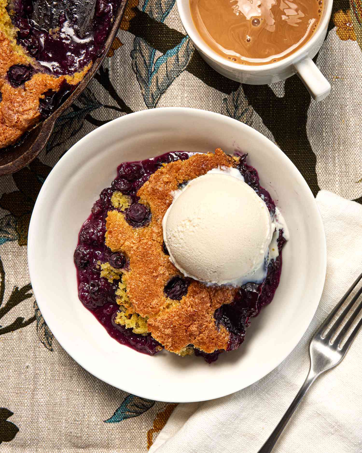Blueberry cornbread cobbler topped with ice cream served on a table with a cup of coffee and a fork beside it