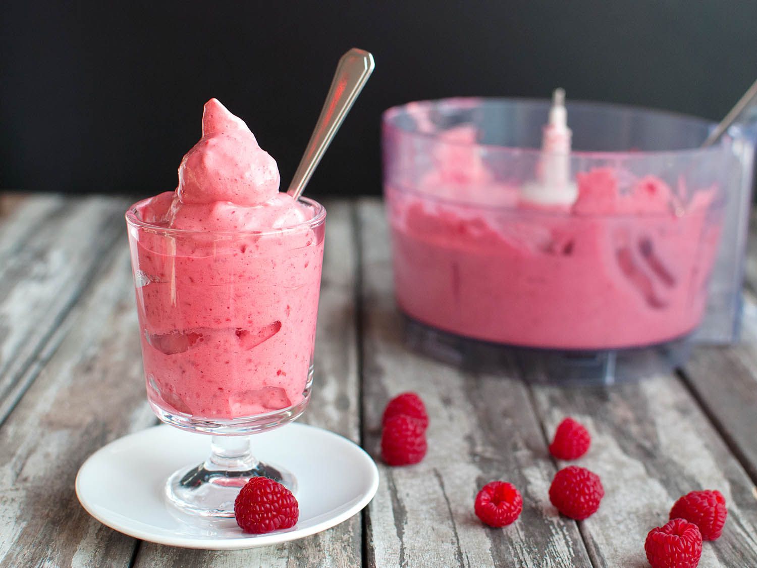 A parfait glass of five-minute fruit mousse, with a food processor bowl containing more mousse in the background. A few raspberries are on the table around the parfait glass.