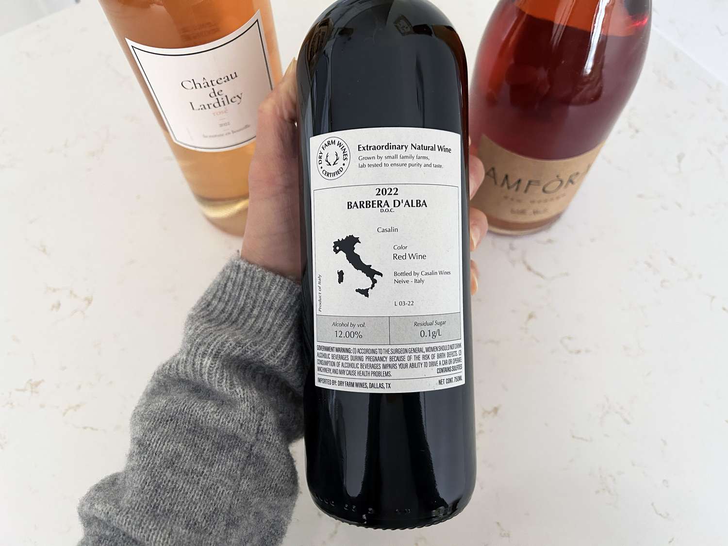 a hand holding a bottle of natural wine against a granite countertop background