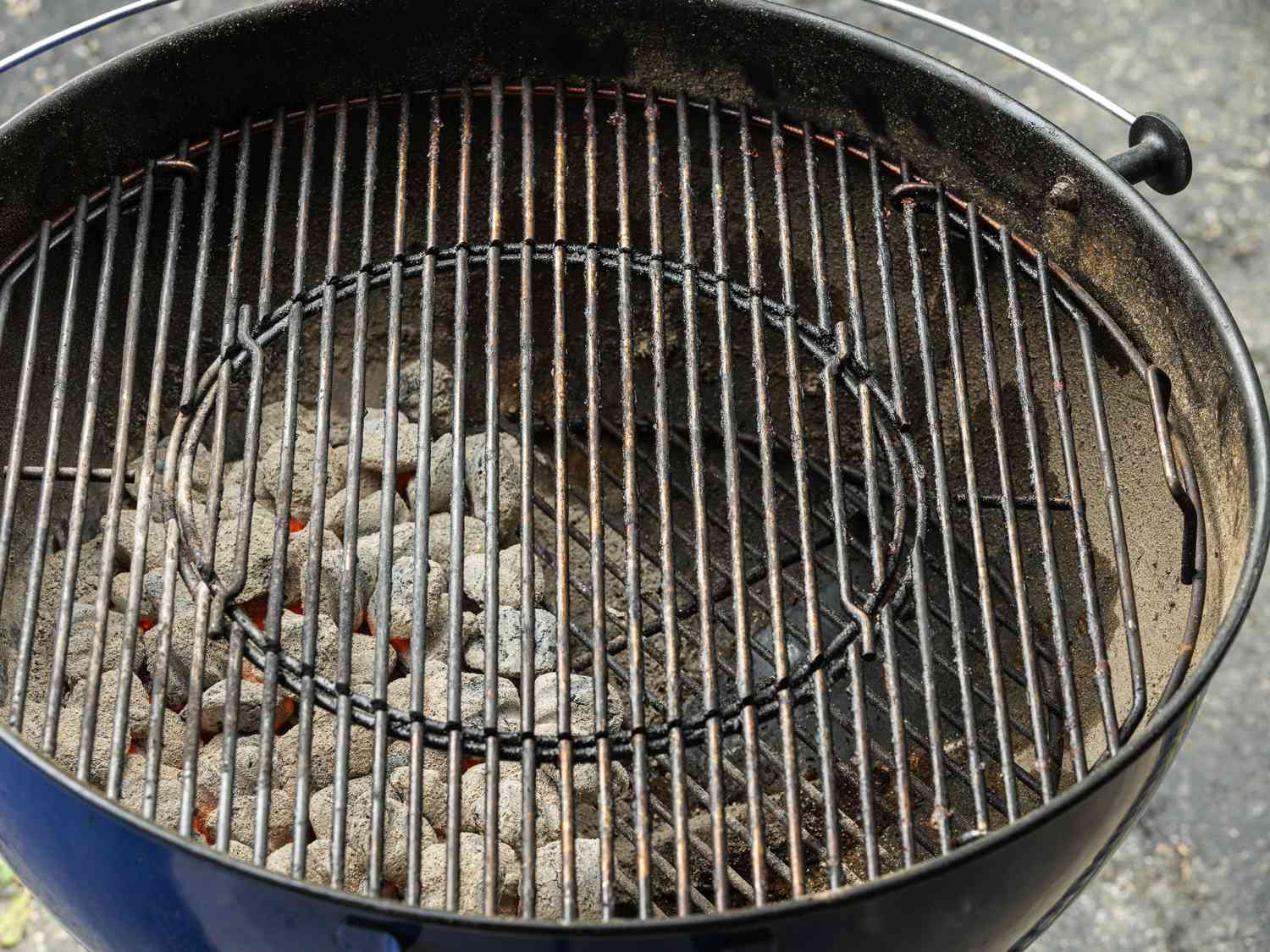 An angled overhead shot of a kettle grill holding charcoal. The lefthand side of the grilling basin is lined with hot, ash-grey coals, while the right side is bare.