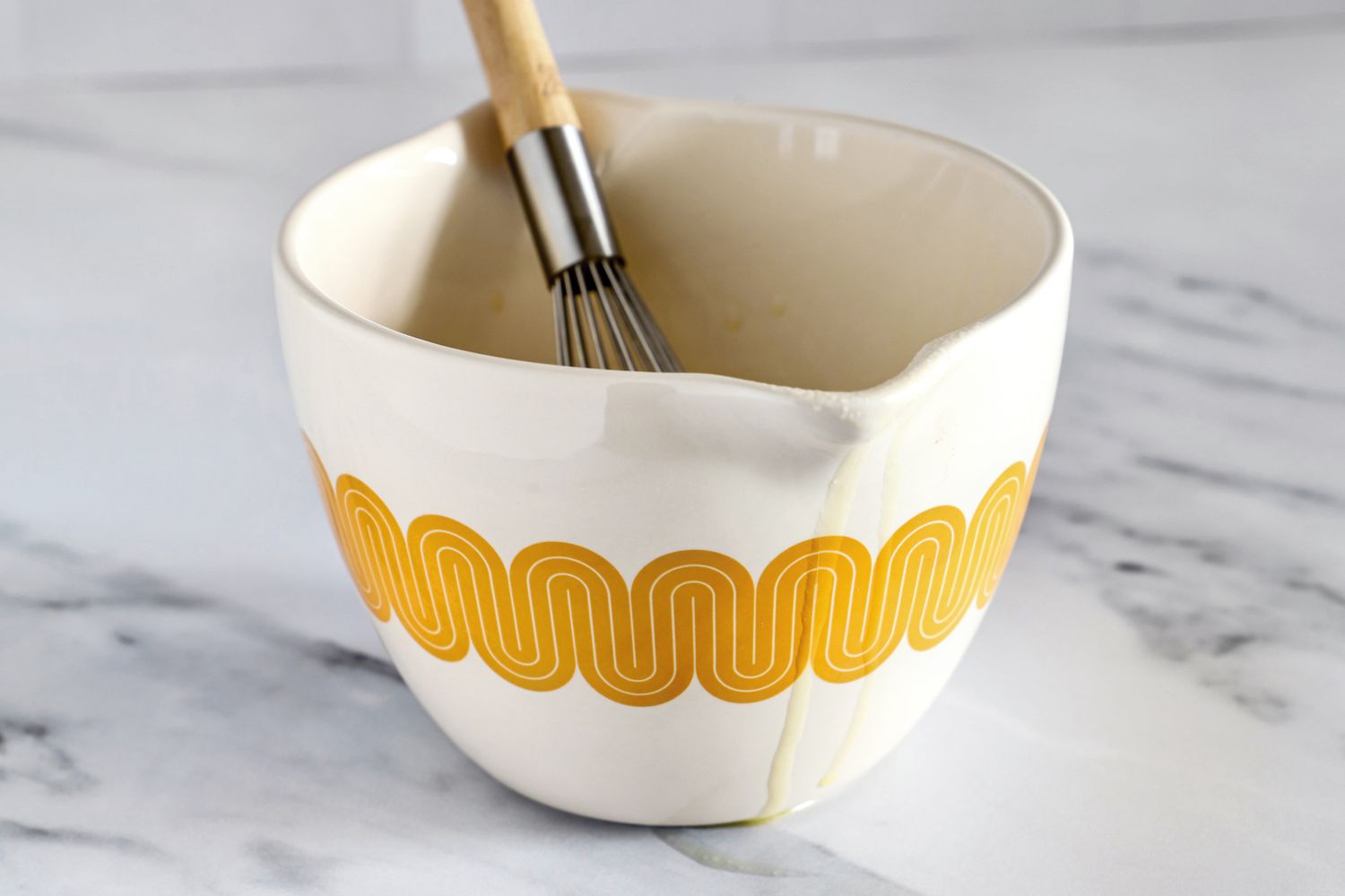 A Great Jones Stir Crazy ceramic mixing bowl with a whisk inside placed on a countertop, with drips running down the side of the bowl