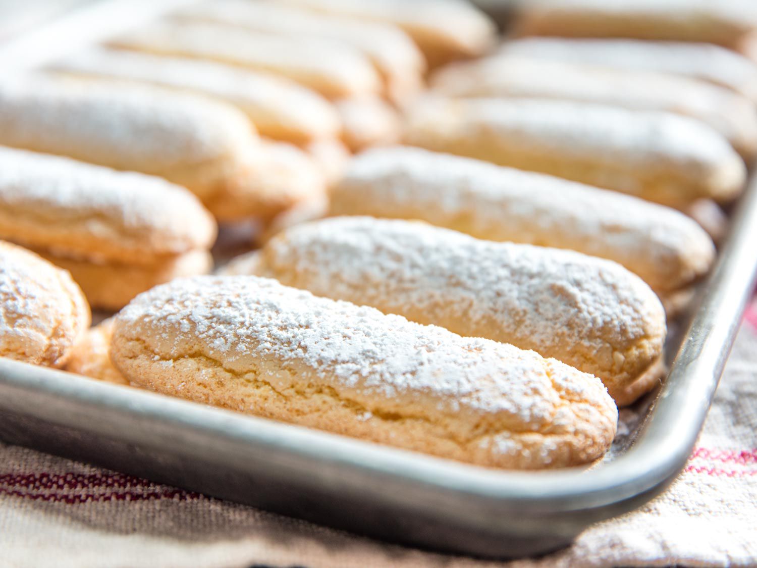Baked homemade ladyfingers on rimmed baking sheet set on red and white striped towel.